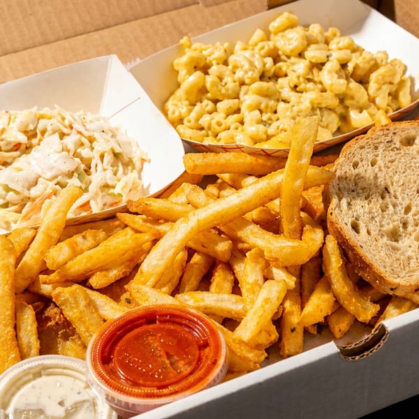 A takeout box filled with french fries, coleslaw, mac and cheese, a slice of bread, and dipping sauces.